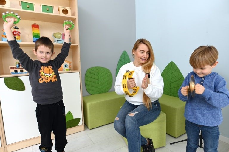 Dvaja chlapci spolu s učiteľkou hrajú na detských hudobných nástrojoch v triede. Jeden chlapec drží dve tamburíny nad hlavou, druhý hrá na čineloch, a učiteľka drží tamburínu, pričom spieva alebo tlieska. V pozadí sú drevené regály s hračkami a zelené dekorácie v tvare listov.
