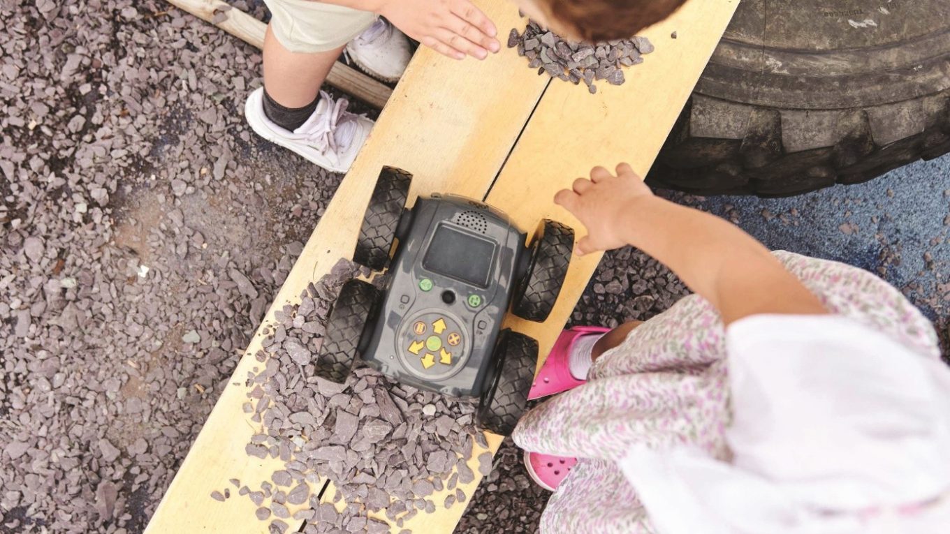 Skupina dětí se zabývá zkoumáním terénního robota na pískovišti, přičemž se učí o technologiích a spolupráci při ovládání robota.