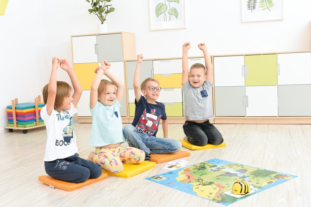 Deti s radosťou sedia na podložkách a dvíhajú ruky pri hre s robotickou včielkou Bee-Bot na vzdelávacej mape
