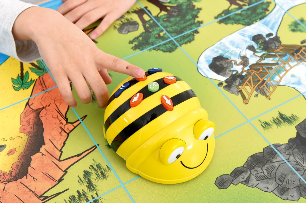 Detail ruky dieťaťa, ktoré stláča tlačidlá na robotickej včielke Bee-Bot na farebnej mape.
