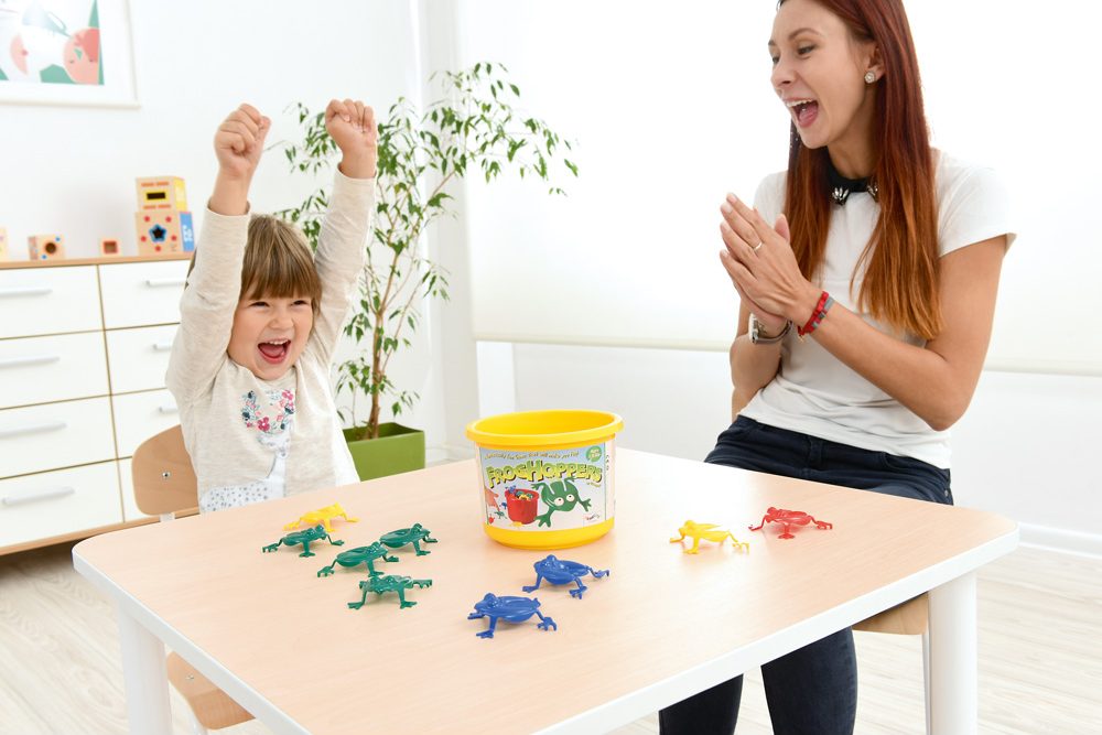 Dieťa a dospelá osoba sa hrajú so skákajúcimi žabkami pri stole v herni.
