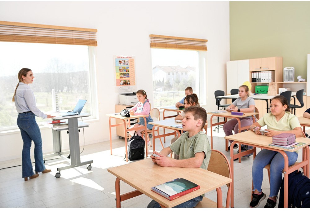 Učiteľka vedie vyučovanie v modernej triede, kde žiaci sedia pri školských laviciach.